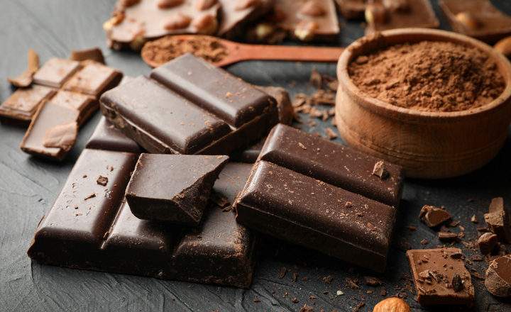 A high-angle, close-up shot of various chocolate products arranged on a dark, textured surface. In the foreground, several thick chunks of dark chocolate are stacked, showing smooth surfaces and broken edges with fine chocolate dust. To the right, a small wooden bowl is filled with fine cocoa powder. In the background, pieces of milk chocolate with embedded whole nuts are scattered alongside a wooden spoon filled with more cocoa. The warm, low lighting emphasizes the rich brown tones and varied textures of the chocolate, creating an indulgent and decadent feel.