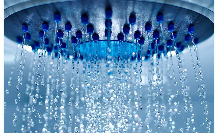 A low-angle, close-up shot of a modern circular showerhead with blue nozzles. Clear streams of water spray downward, breaking into shimmering droplets and fine mist as they fall. The lighting is cool and blue-toned, highlighting the movement and clarity of the water against a soft, out-of-focus background.