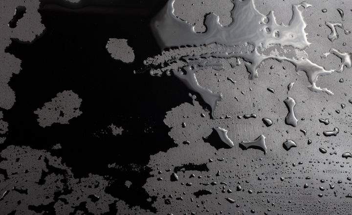 A high-contrast, top-down photograph of water droplets and pooled liquid on a dark, matte surface. On the left, the liquid forms thin, jagged, lace-like patterns against the black background. On the right, larger, more cohesive pools and individual droplets catch the light, revealing a smooth, reflective texture. The lighting creates a dramatic effect, emphasizing the contrast between the wet and dry areas of the surface.