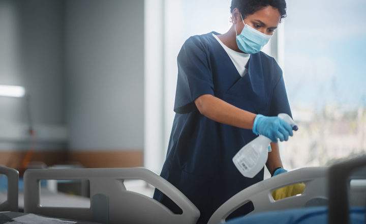 A healthcare worker wearing a navy blue scrub top, a light blue surgical mask, and blue nitrile gloves is shown cleaning a hospital room. They are leaning over a hospital bed, using a white spray bottle to apply a cleaning agent to the plastic bed frame while holding a yellow cloth in their other hand. The background is a brightly lit, clean clinical environment with a large window. The focus is on the worker's hands and the cleaning process, emphasizing hygiene and sanitization protocols.
