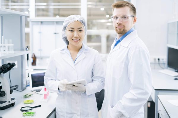 Cheerful young scientists in laboratory coats standing at desks with food samples and equipment holding tablet and looking at camera smiling